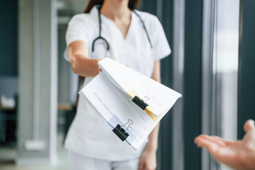 Healthcare professional in white scrubs extending an arm with stacked forms clipped in a doorway area, offering paperwork to a visitor